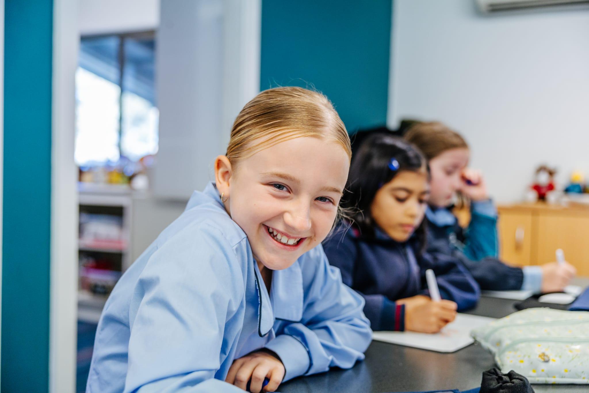 Primary student smiling at her desk while classmates write in class
