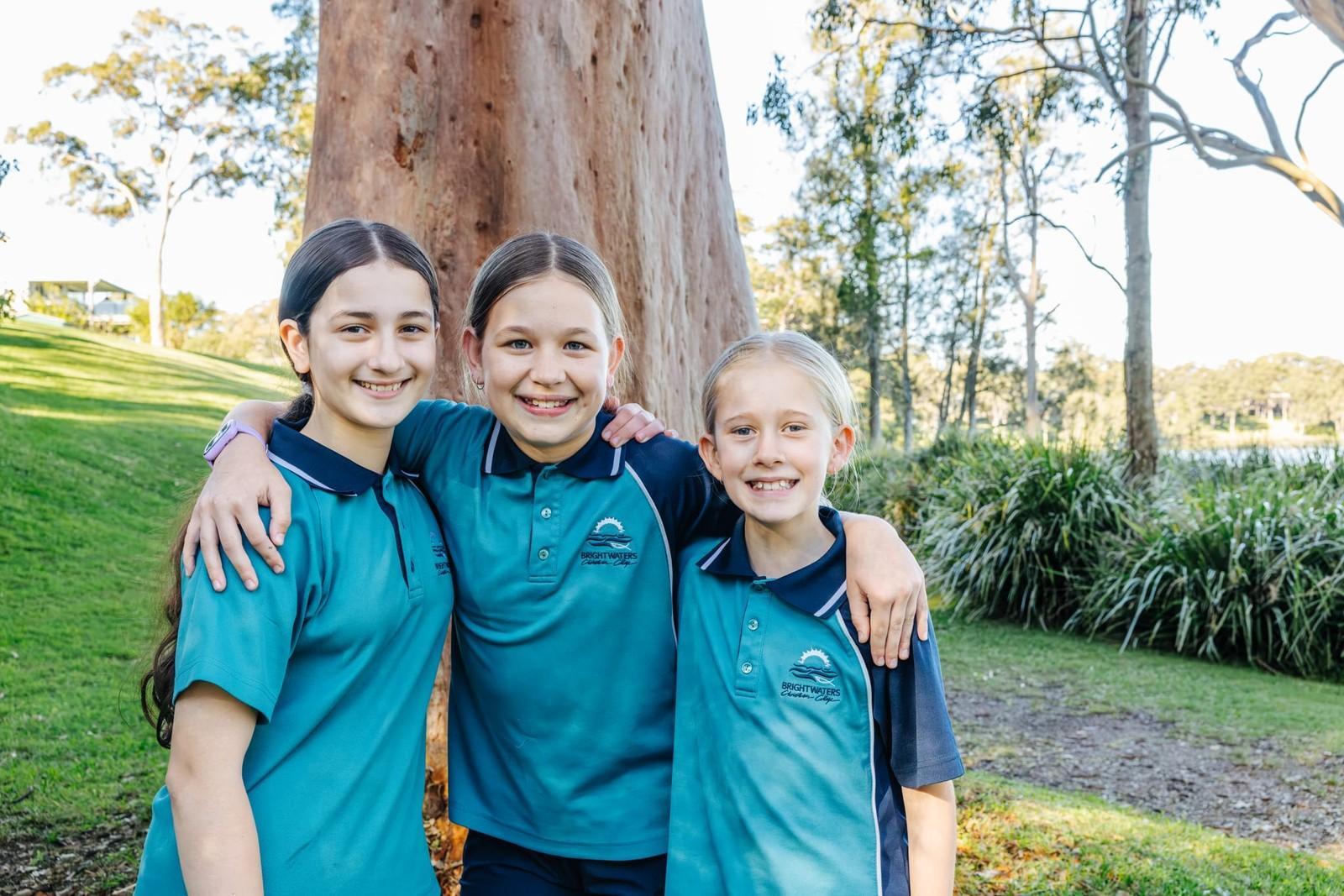 Three smiling primary students arm in arm under gum trees on the school grounds.