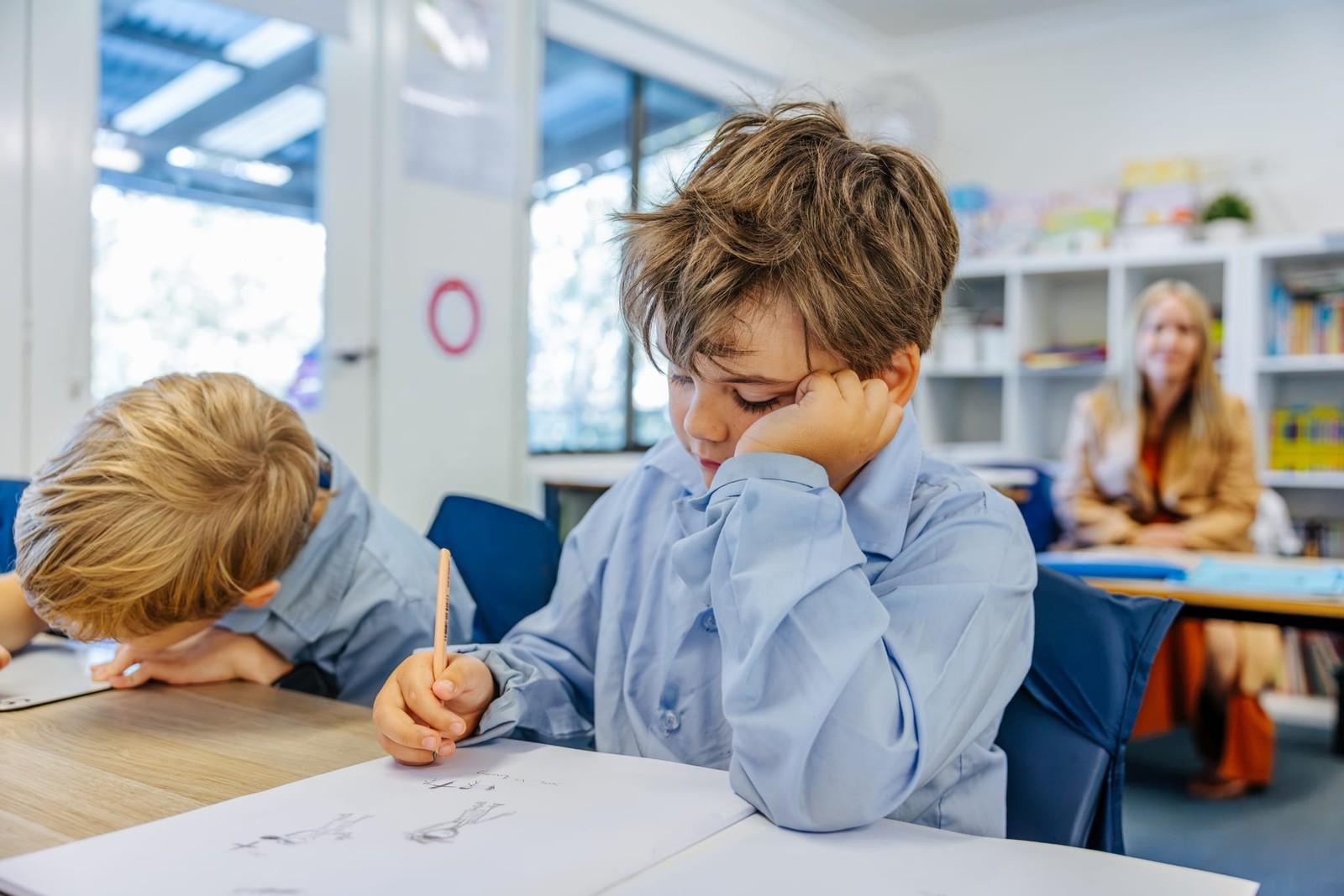 Young student concentrating on his drawing at a classroom desk while his teacher looks on