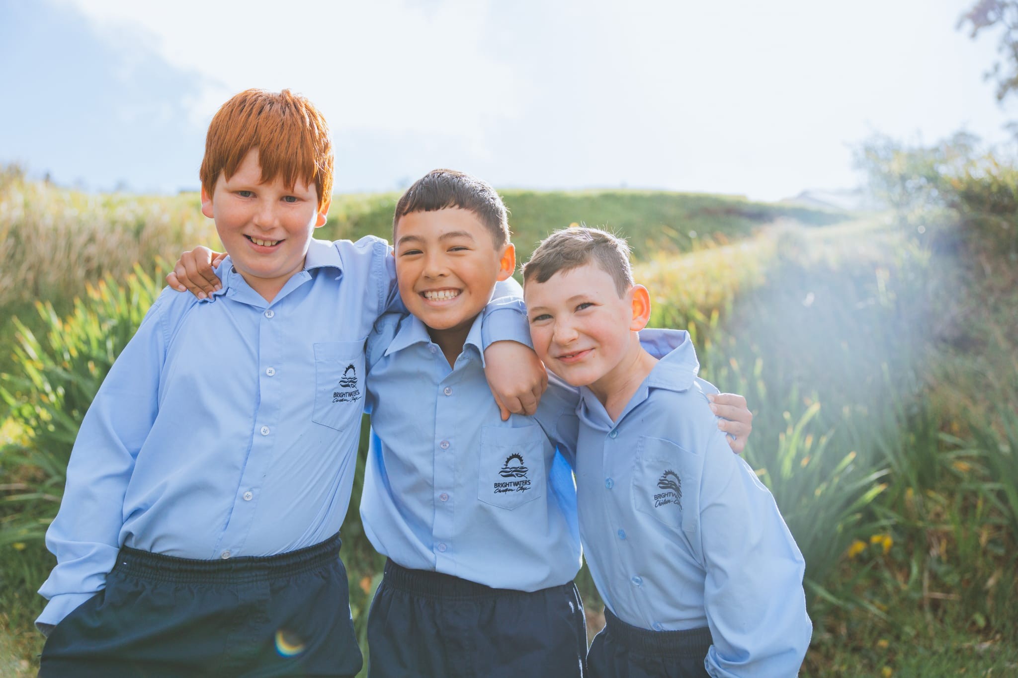 Three smiling Brightwaters Christian College students in light-blue uniforms with arms around each other, standing outdoors in sunlit grass